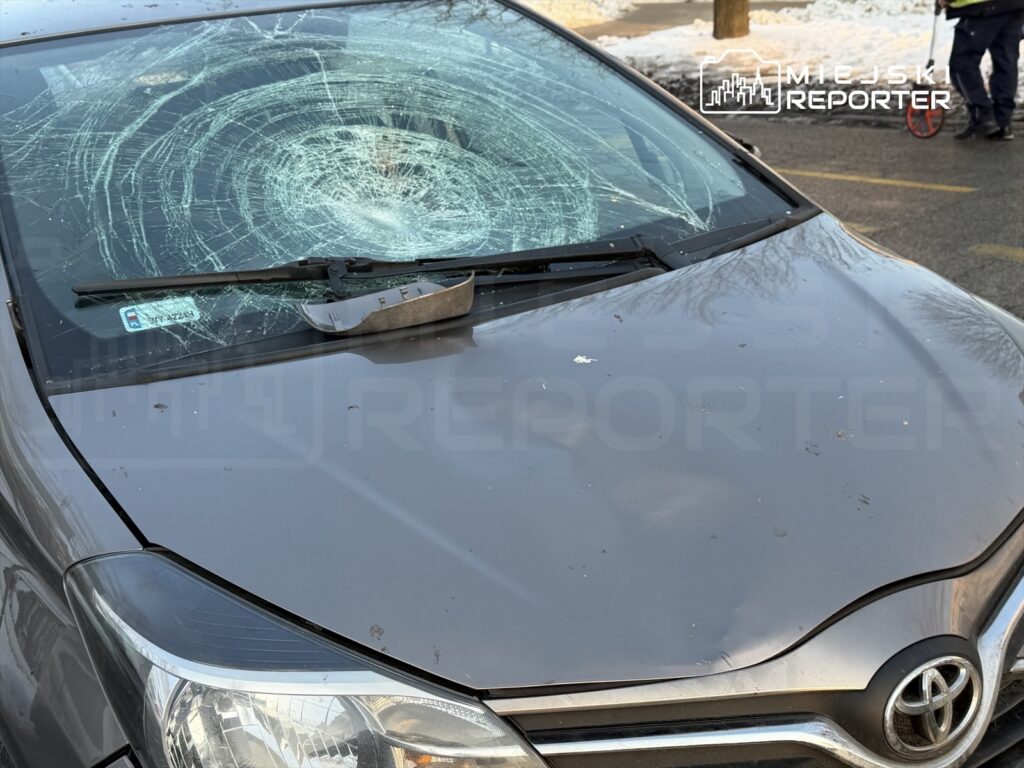 Pęknięta szyba czołowa szarego samochodu osobowego, na której widoczne są ślady uderzenia. W tle widać postać stojącą obok pojazdu.