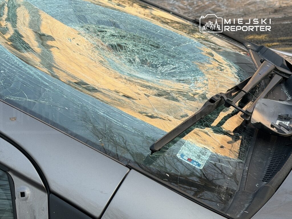 Zniszczona szyba czołowa samochodu z pęknięciami w kształcie spirali, leżąca na parkingu.