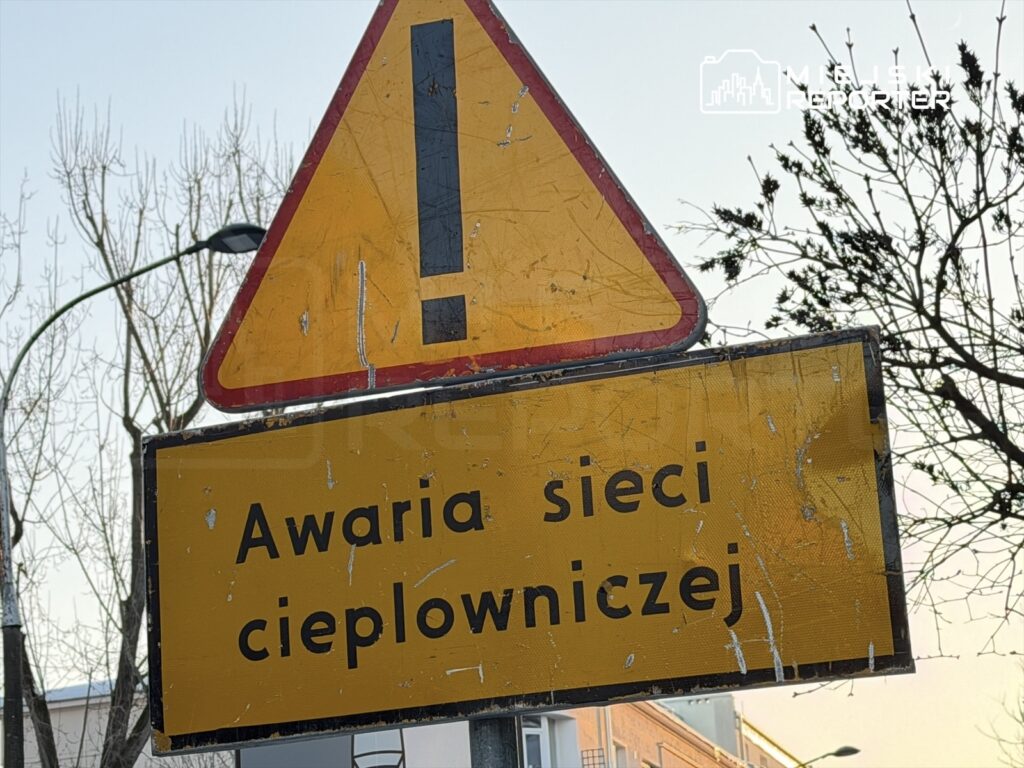 Żółty znak ostrzegawczy informujący o awarii sieci ciepłowniczej, w tle gołe drzewa i budynki.