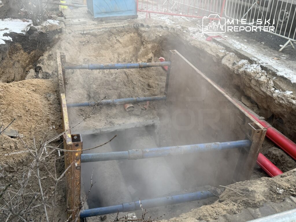 Wykop na budowie z niebieskimi i czerwonymi rurami, otoczony przez piasek i dym, z zabezpieczeniami stalowymi.