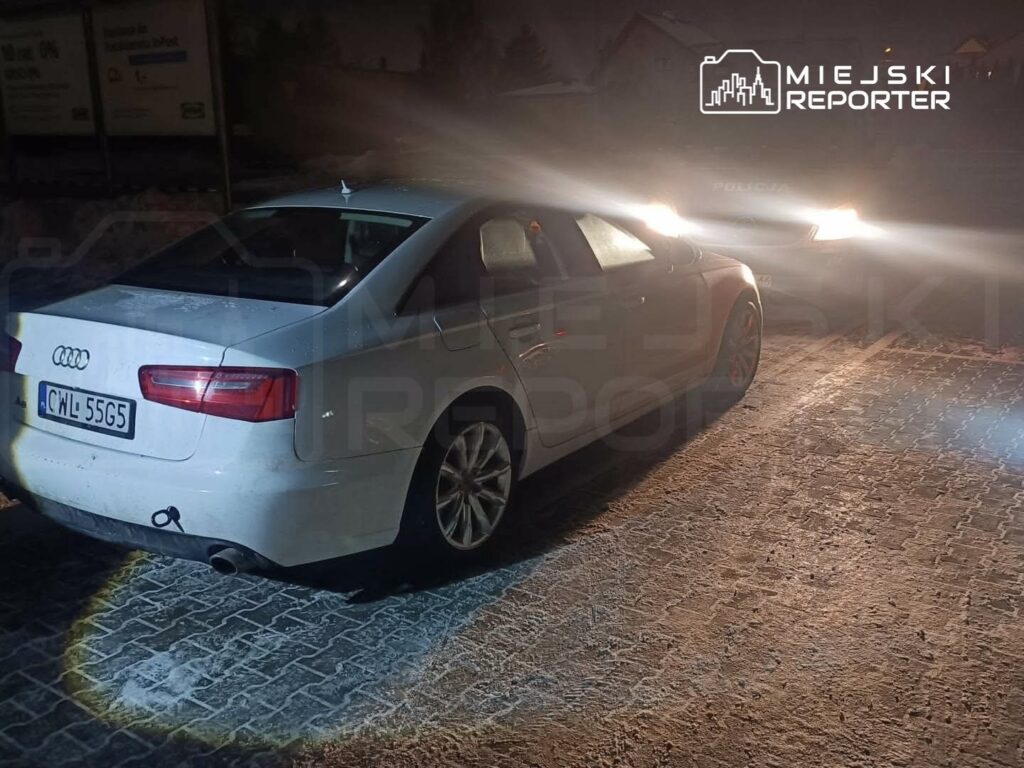 Białe Audi zaparkowane na oświetlonym chodniku, w tle radiowóz policyjny z włączonymi światłami.