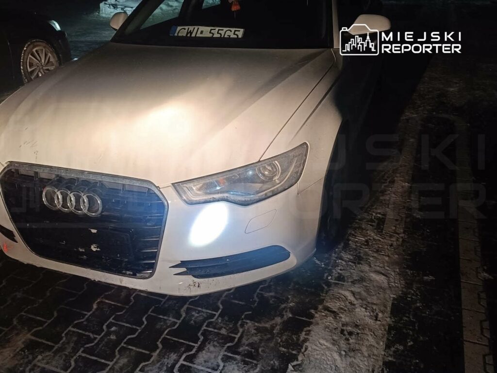 Biały samochód osobowy marki Audi zaparkowany na oświetlonym, zimowym chodniku.