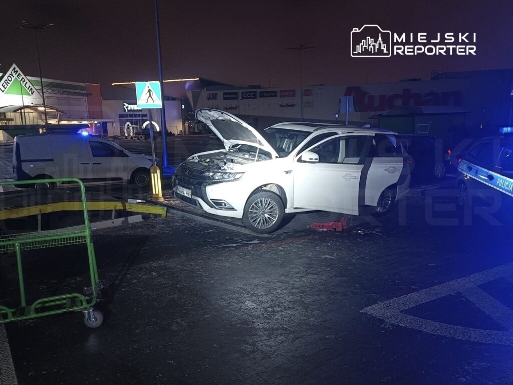 Biały samochód osobowy z otwartą maską stoi na parkingu, obok zaparkowany radiowóz policyjny.