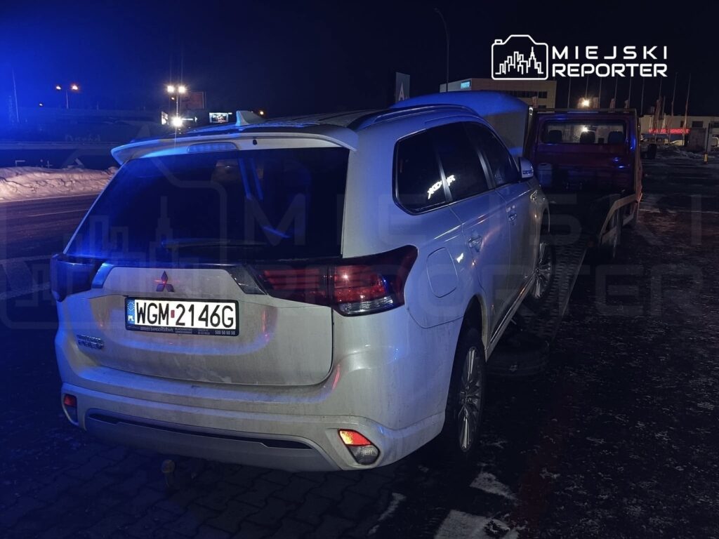 Biały SUV z tablicą rejestracyjną WGM-2146G zaparkowany na drodze, oświetlony niebieskimi światłami policyjnymi.