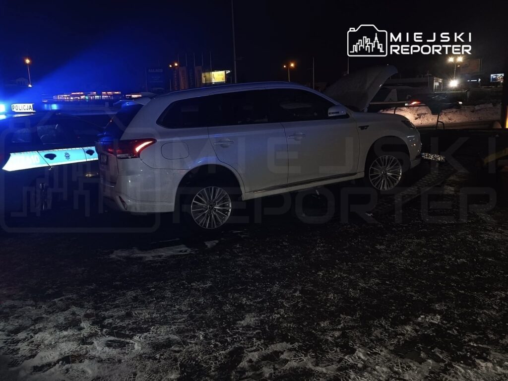 Biały samochód osobowy z otwartą maską stoi obok radiowozu policyjnego z włączonymi sygnałami świetlnymi.