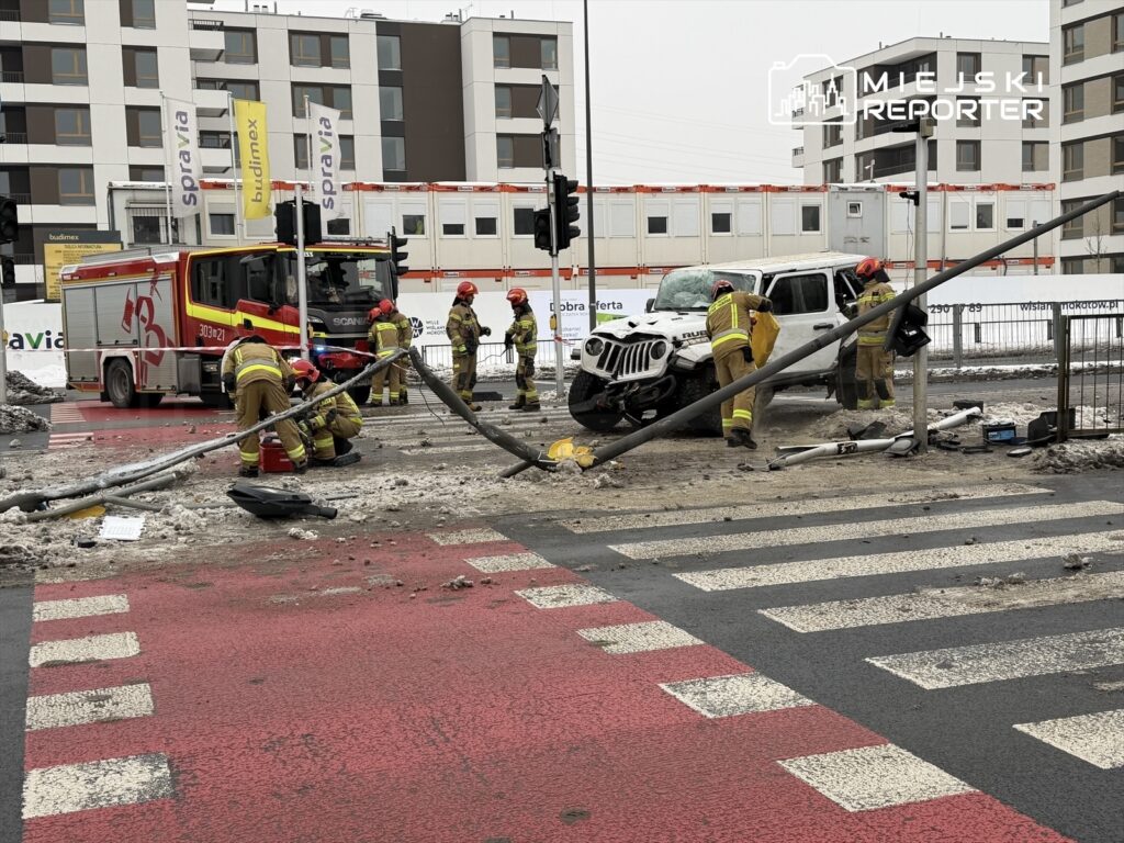 Strażacy w kaskach i mundurach pracują przy uszkodzonym samochodzie, obok wozu strażackiego na skrzyżowaniu.