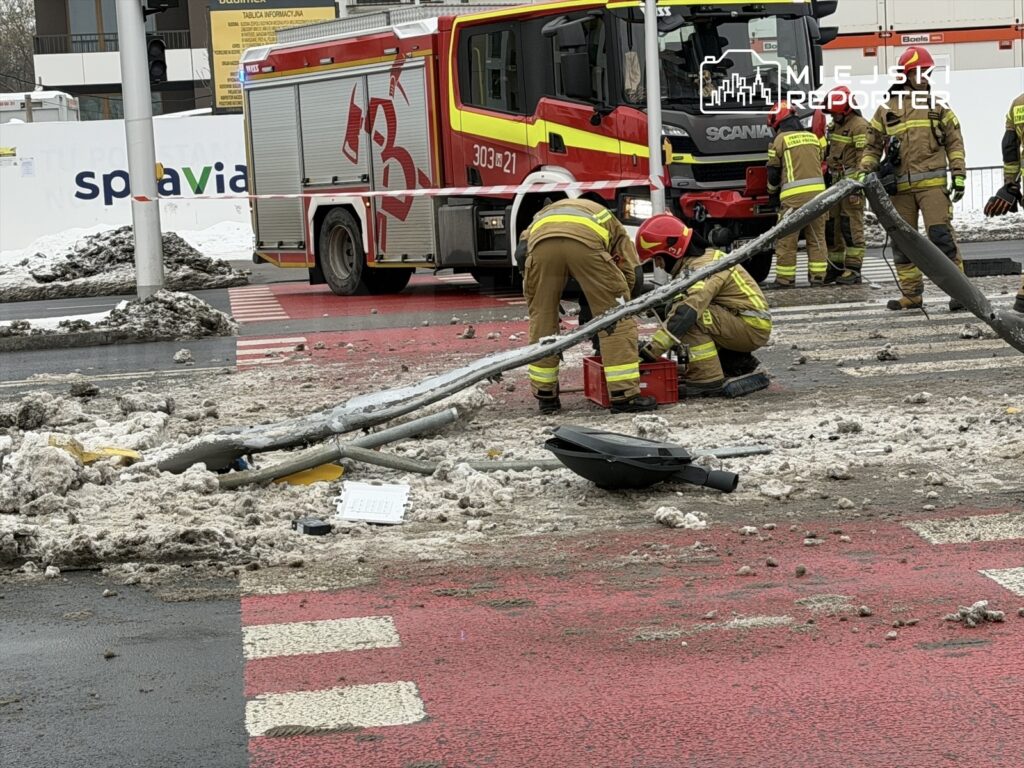 Strażacy w mundurach pracują przy uszkodzonym słupie elektrycznym na zaśnieżonej jezdni.