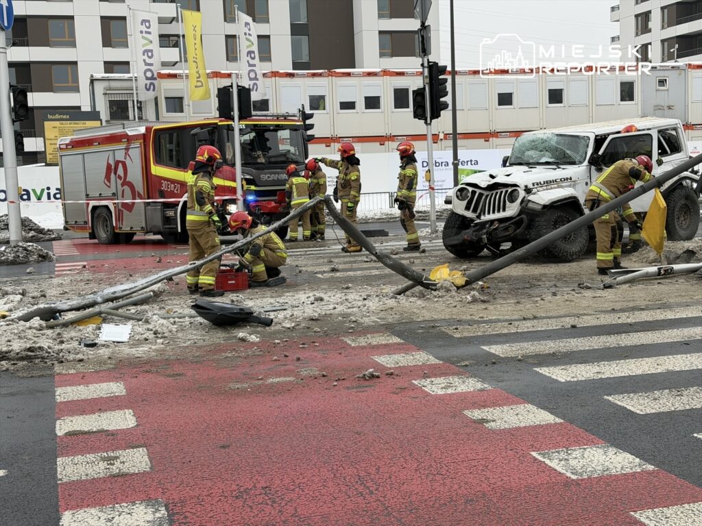 Grupa strażaków w mundurach pracuje przy wypadku drogowym, otaczając uszkodzony samochód i przewrócone słupy.