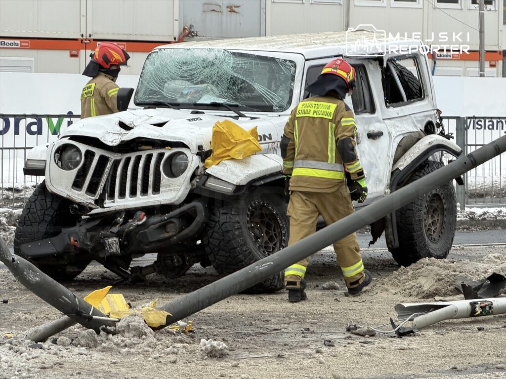 Uszkodzony biały samochód terenowy stoi na drodze, obok niego strażacy w mundurach zabezpieczających przeszukują okolicę.