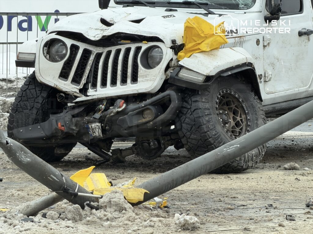 Uszkodzony biały samochód terenowy z rozbitym przodem leży na zaśnieżonej nawierzchni obok uszkodzonego słupa.