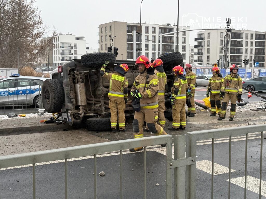 Grupa strażaków w hełmach, pracująca przy przewróconym pojeździe na ruchliwej ulicy w mieście. W tle widoczne budynki mieszkalne i radiowóz policyjny.