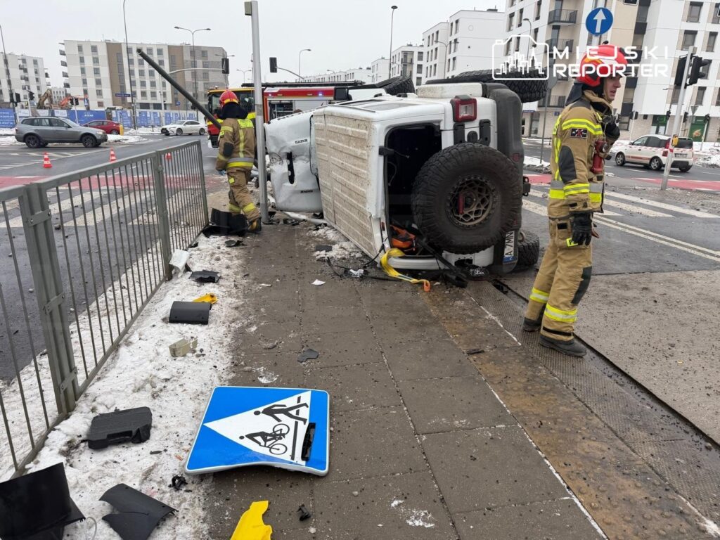 Strażacy podnoszą przewrócony pojazd na ulicy, obok uszkodzone znaki drogowe i fragmenty asfaltu.