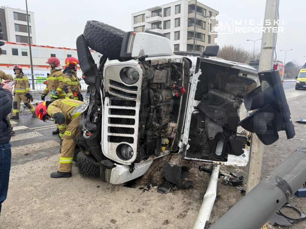 Na drodze leży przewrócony samochód terenowy, wokół niego pracują strażacy w hełmach, a w tle widać nowoczesne budynki.
