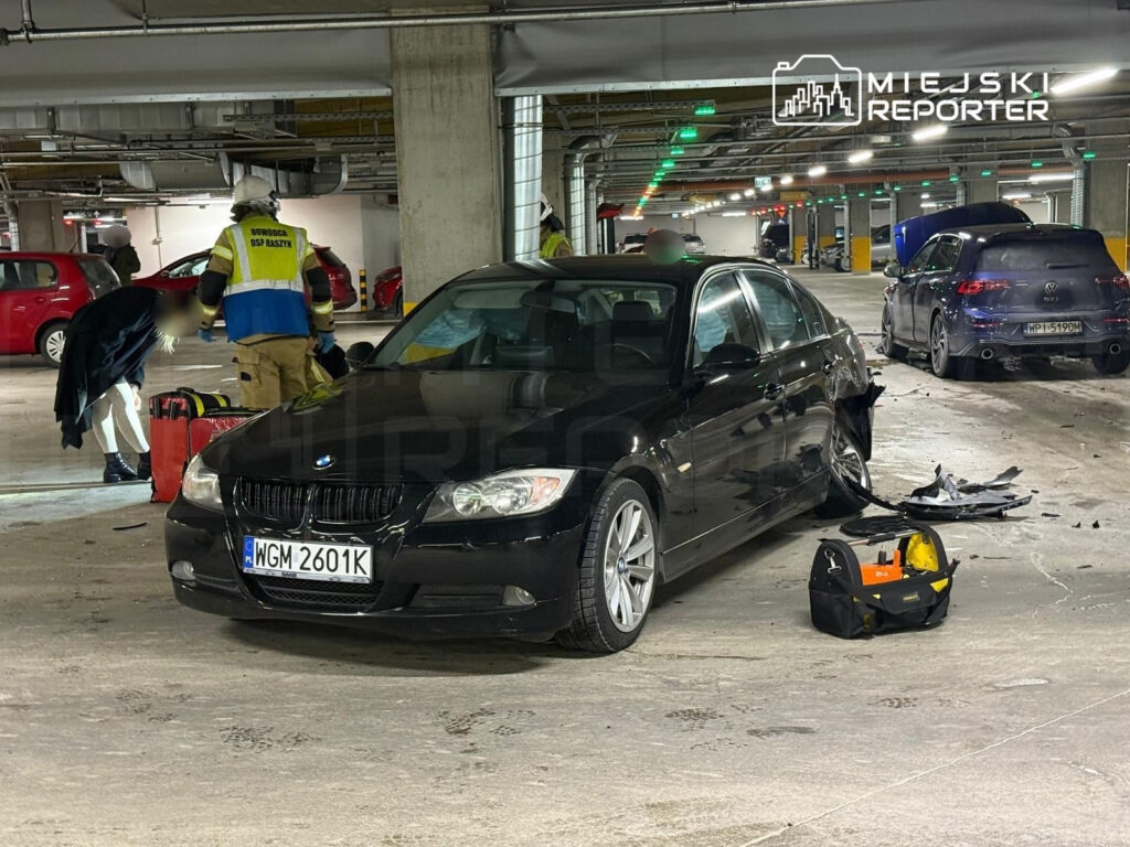 Ratownicy udzielają pomocy poszkodowanemu przy czarnym samochodzie w podziemnym parkingu. W tle widać inne pojazdy.