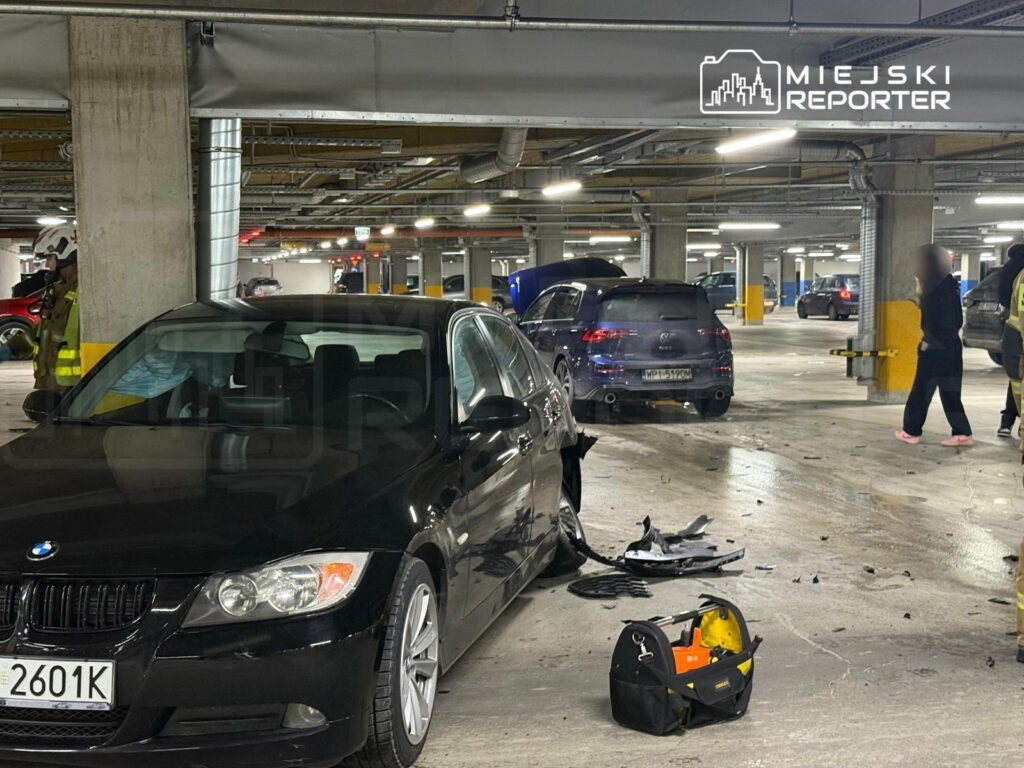 Czarny samochód osobowy z uszkodzeniami stoi na parkingu, obok pracują strażacy w kamizelkach odblaskowych.