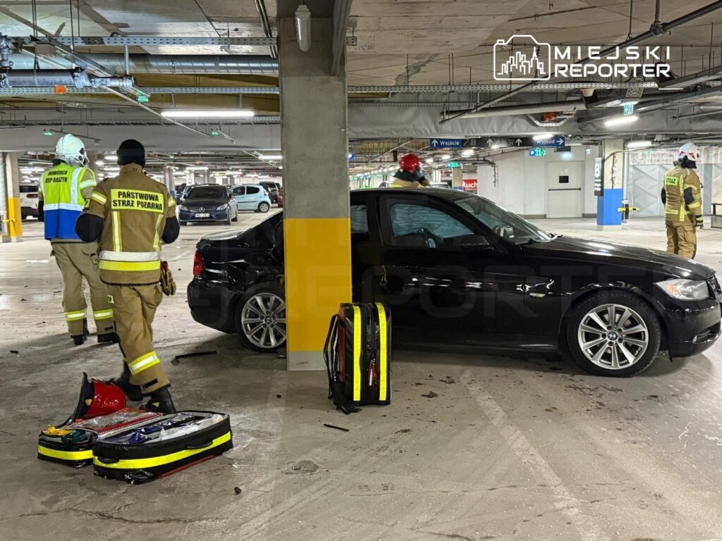 Strażacy w mundurach pracują w podziemnym parkingu, obok czarnego samochodu, z narzędziami ratunkowymi na ziemi.