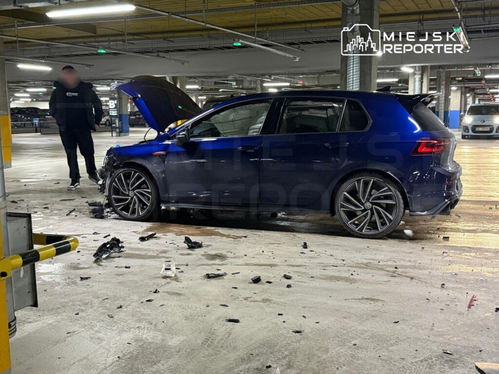 Niebieski samochód osobowy z uszkodzoną przednią częścią stoi na parkingu, obok mężczyzna w czarnej kurtce.