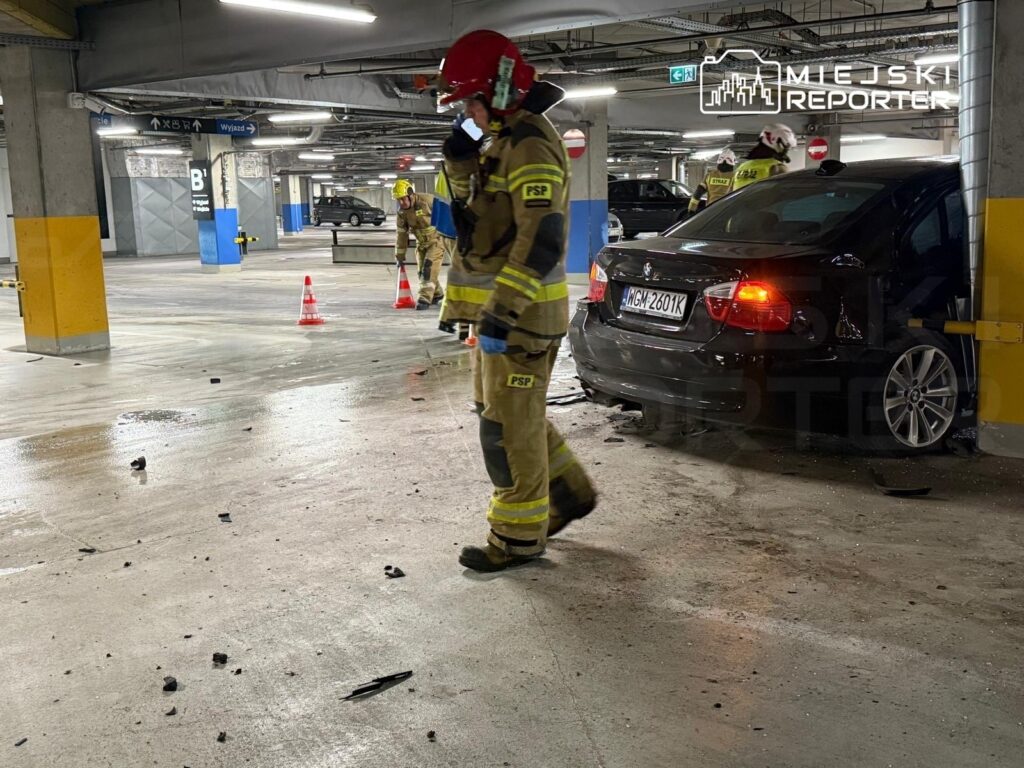 Strażak w hełmie i mundurze przemieszcza się po parkingu podziemnym, obok uszkodzonego samochodu.