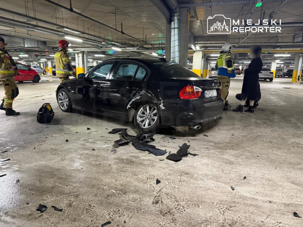 Czarny samochód osobowy z uszkodzeniami stoi na parkingu, obok pracują strażacy w mundurach, a także kobieta w czarnym płaszczu.