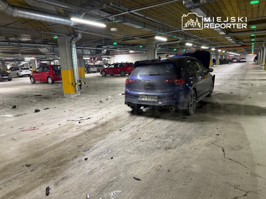 Czarny samochód osobowy z otwartą maską stoi na parkingu, wokół leżą fragmenty plastików i innych odpadków.