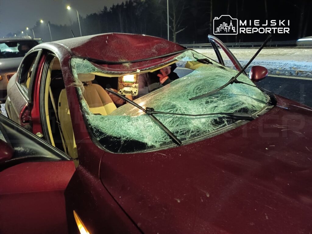 Czerwony samochód z uszkodzoną szybą czołową stoi na drodze nocą, obok widać osobę z lampą.