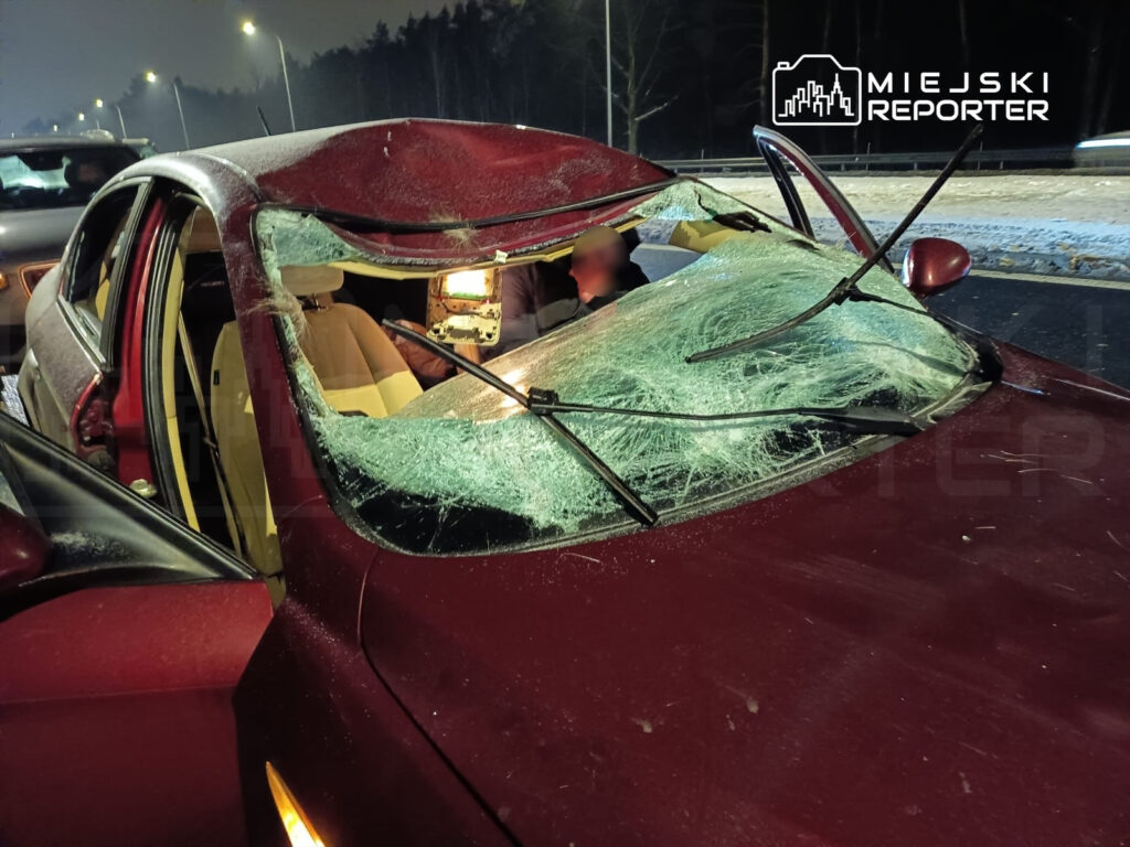 Czerwony samochód osobowy z rozbitym przednim szybą stoi na drodze, a w tle widoczne są inne pojazdy.