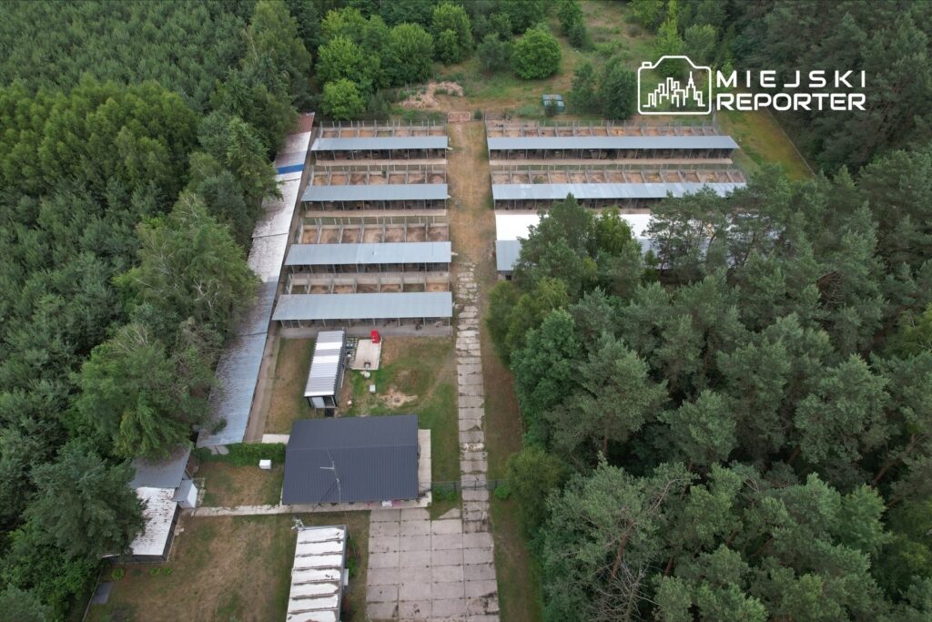 Widok z lotu ptaka na kompleks budynków z metalowymi dachami otoczony zielenią drzew. W centrum widoczna ścieżka.