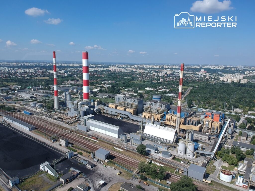 Widok na zakład przemysłowy z czerwono-białymi kominami oraz infrastrukturą kolejową, otoczony zielenią.