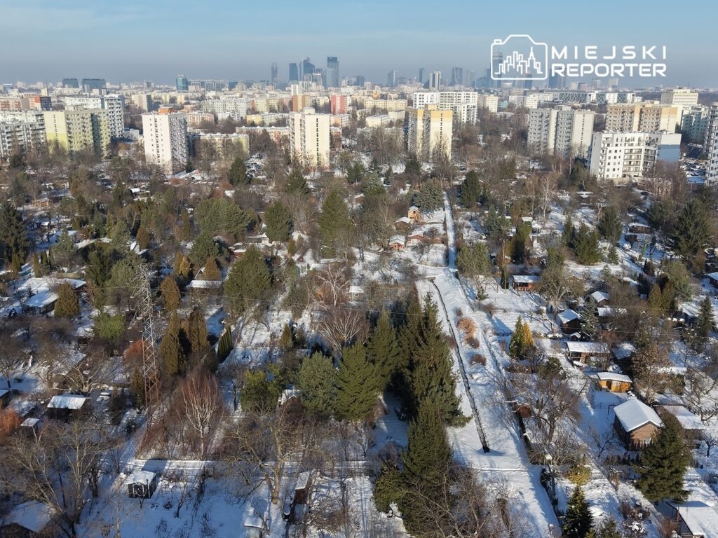 Widok na osiedle z domkami jednorodzinnymi i działkami, pokrytymi śniegiem, w tle nowoczesne wieżowce miejskie.
