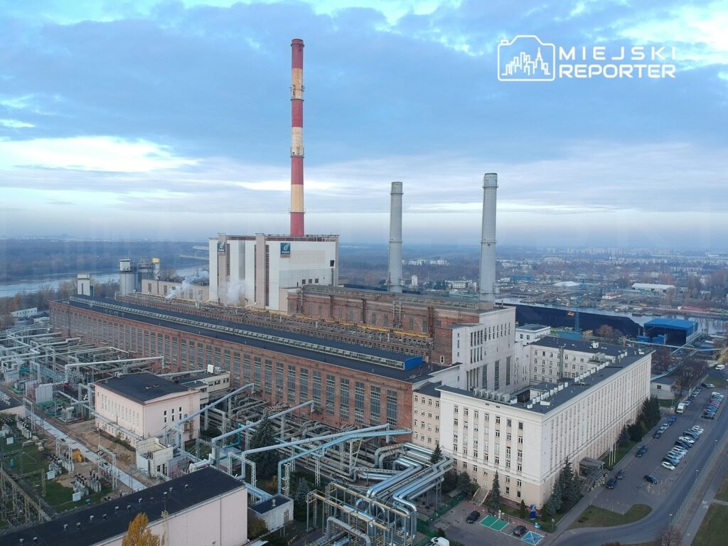 Fabryka przemysłowa z dużymi kominami i instalacjami, otoczona zielenią i parkingiem, w tle widoczna rzeka.