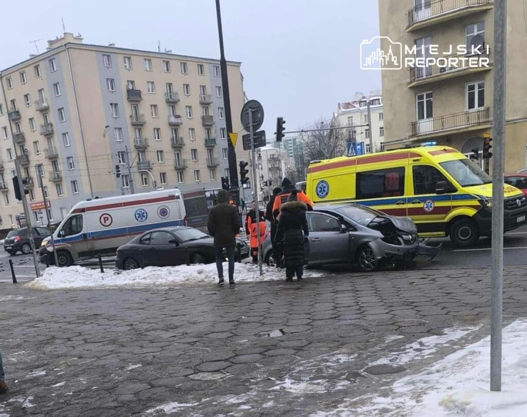 Karetka i strażacy pomagają poszkodowanym po wypadku drogowym w mieście, w tle widać uszkodzony samochód.