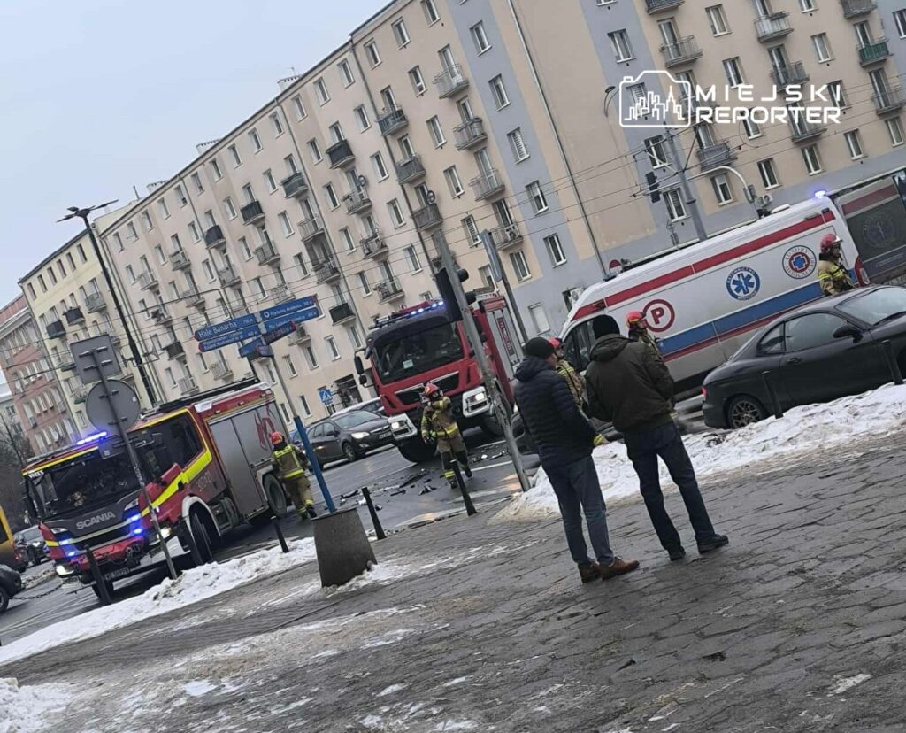 Na ulicy stoi dwóch mężczyzn, obserwujących działania straży pożarnej i karetki pogotowia w otoczeniu bloków mieszkalnych.