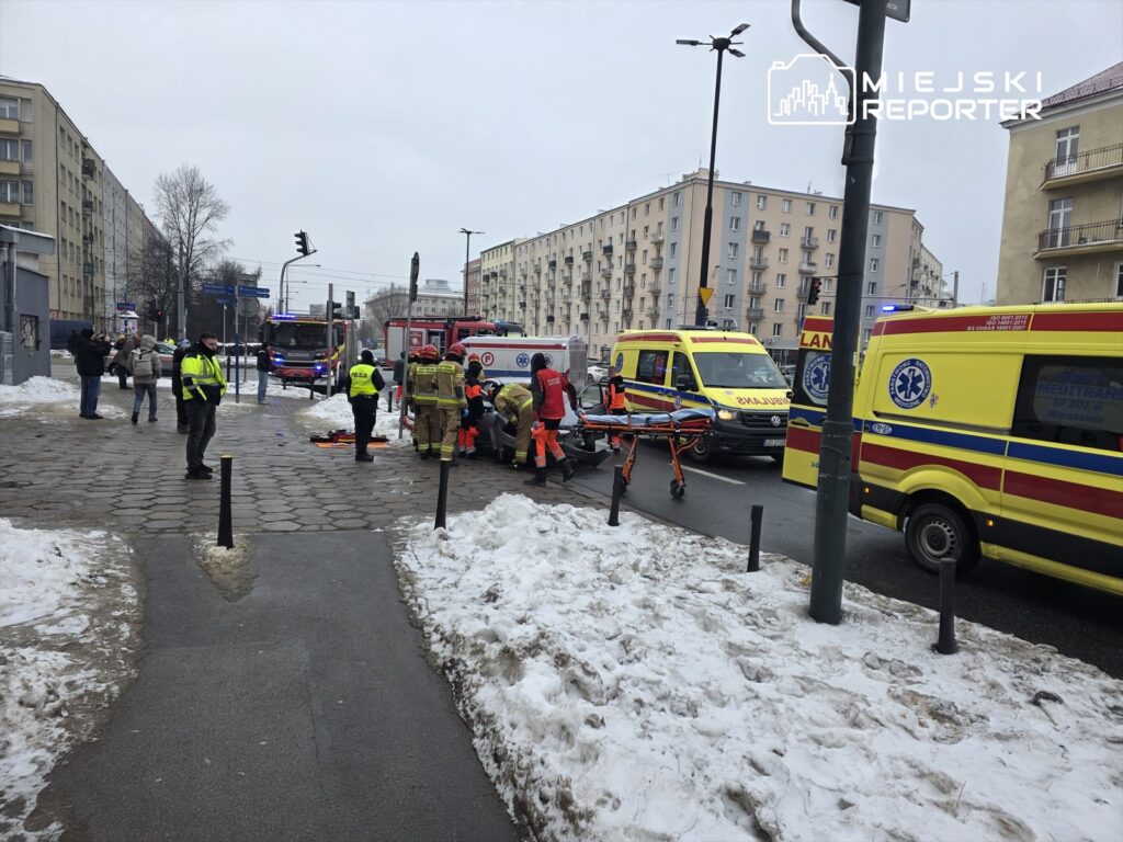 Grupa ratowników medycznych udziela pomocy poszkodowanemu na chodniku, w pobliżu zaparkowanych ambulansów.