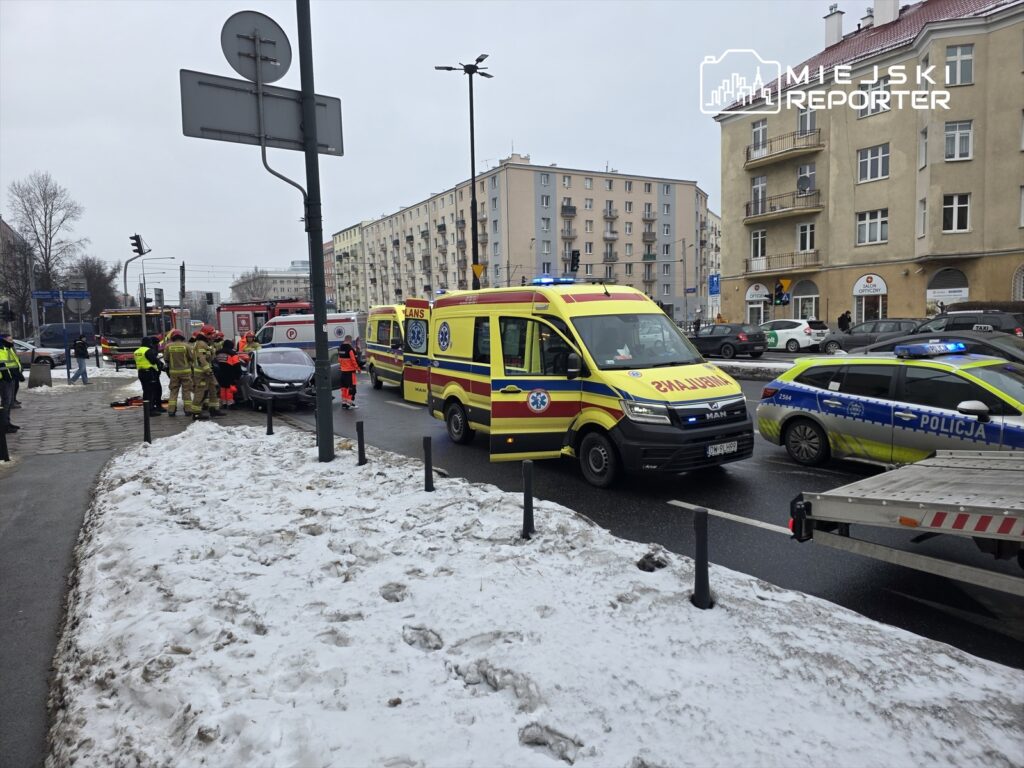 Grupa ratowników w mundurach pomaga poszkodowanemu w wypadku drogowym na ulicy, obok stoją pojazdy służb ratunkowych.