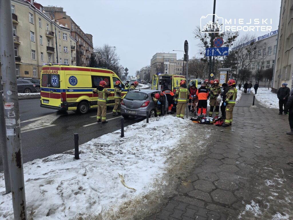 Grupa ratowników medycznych i strażaków udziela pomocy poszkodowanemu przy samochodzie na zaśnieżonej ulicy.