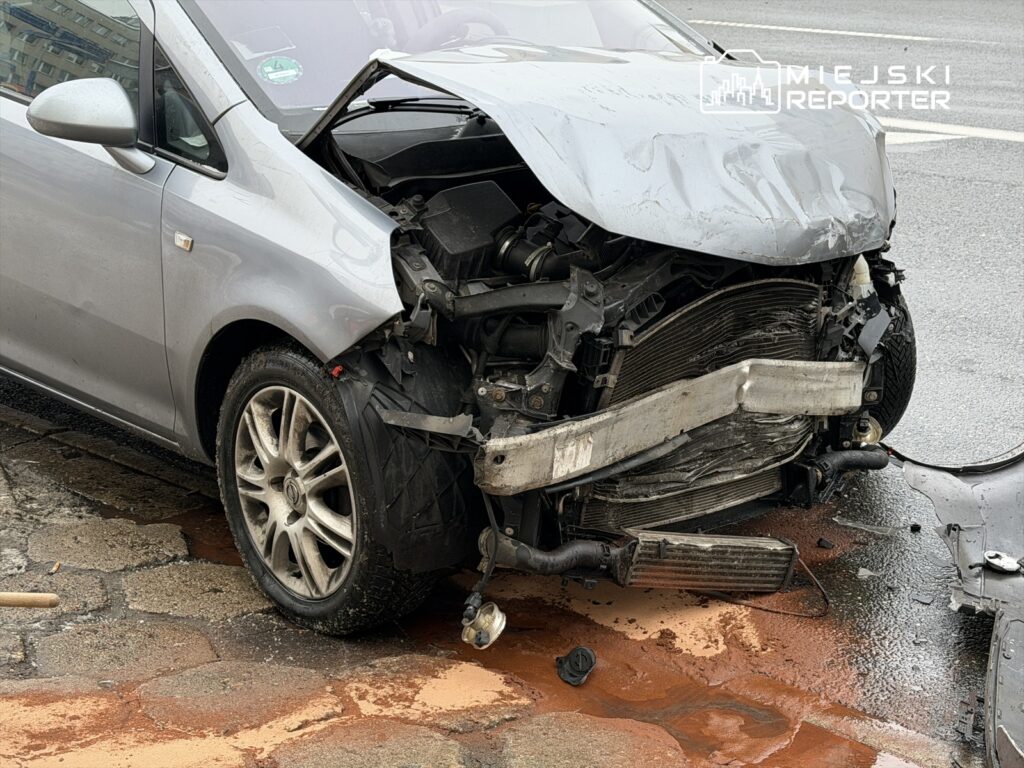Uszkodzony srebrny samochód osobowy z zgniecionym przodem stoi na ulicy, obok widać plamę płynu.