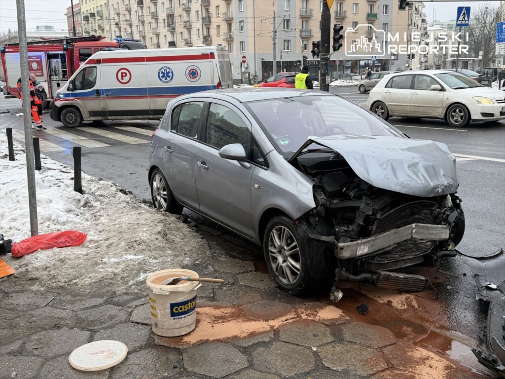 Uszkodzony samochód osobowy stoi przy krawężniku, obok ratownicy medyczni i strażacy interweniują na miejscu wypadku.