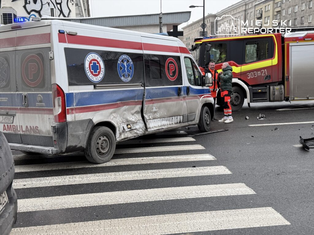 Ambulans z uszkodzeniami stoi na przejściu dla pieszych, obok rozmawiają ratownicy w odzieży ochronnej.