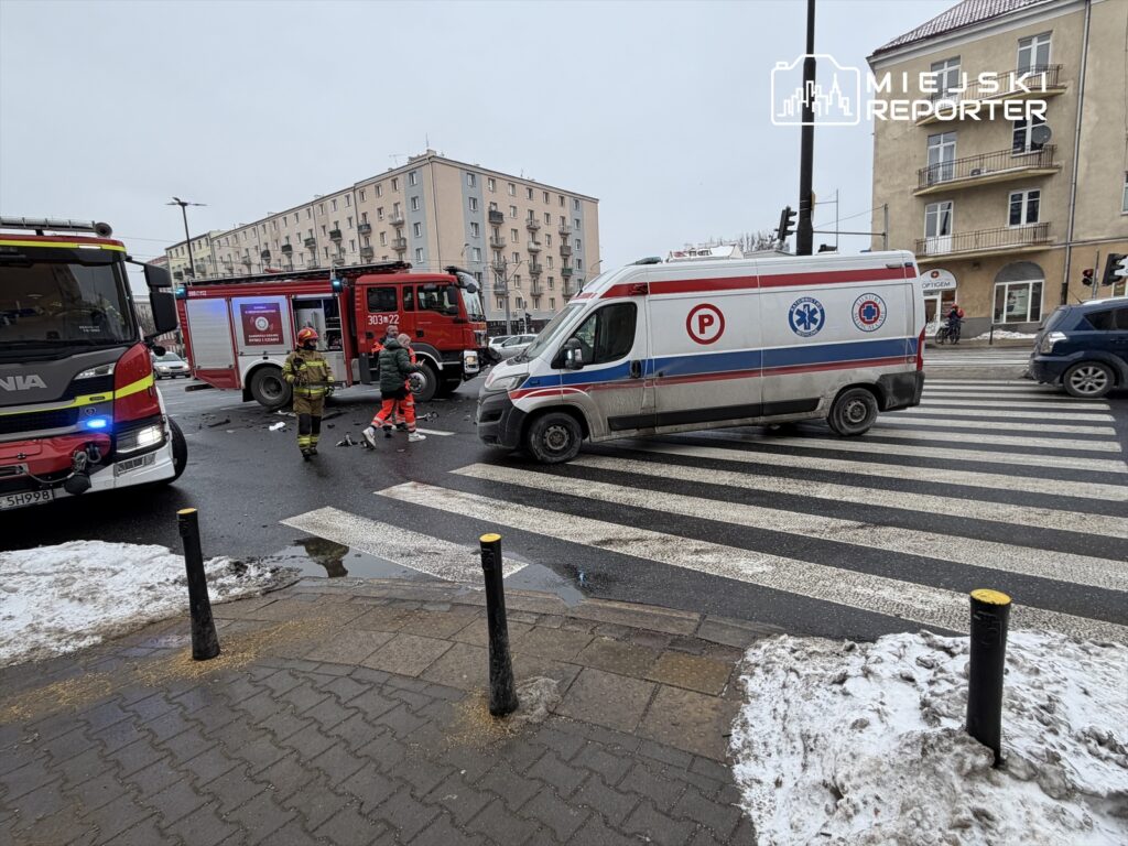 Zespół ratunkowy z ambulansu i strażacy w akcji na skrzyżowaniu, obok pojazdu strażackiego.
