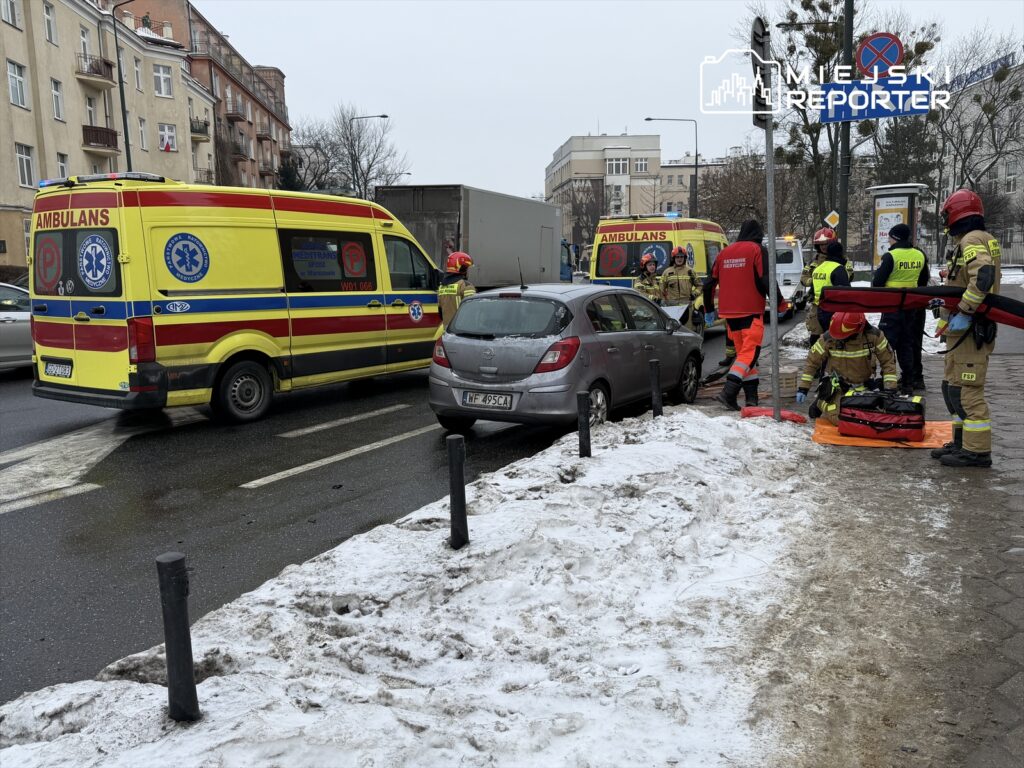 Zespół ratunkowy w akcji, udzielający pomocy poszkodowanemu na chodniku obok zaparkowanych pojazdów.