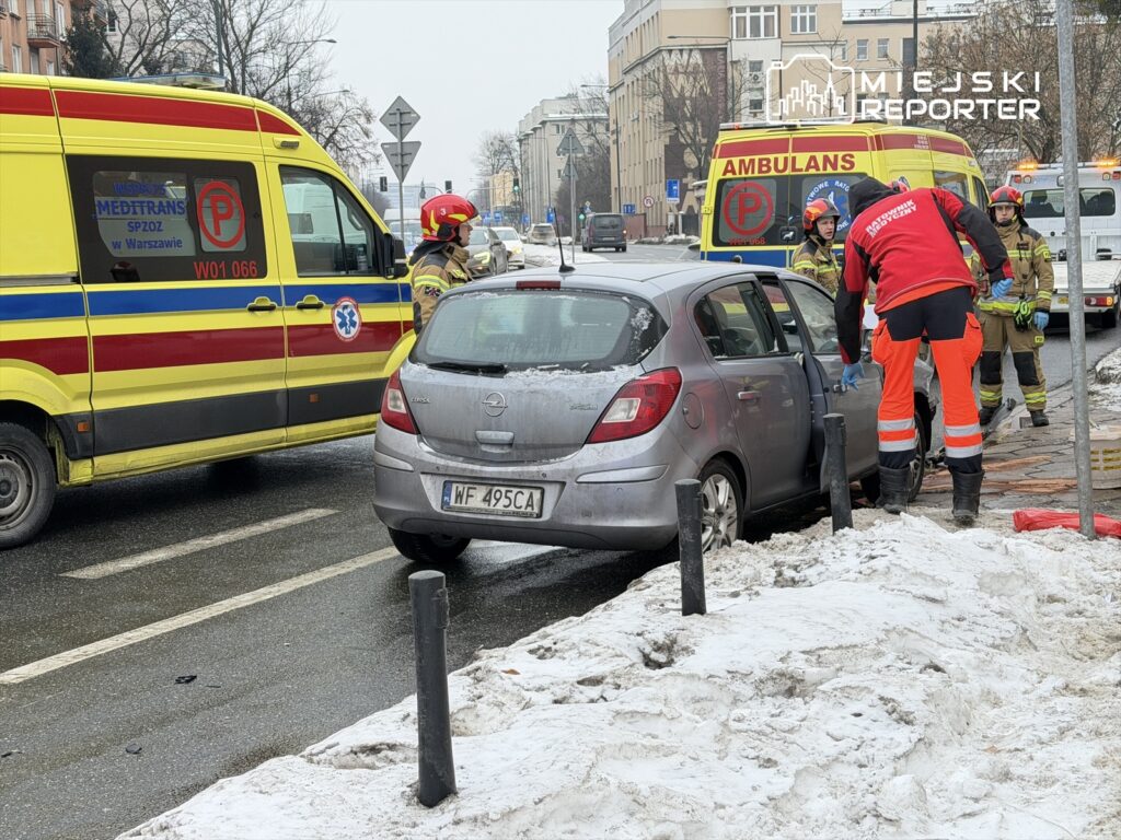 Ratownicy medyczni udzielają pomocy poszkodowanemu w samochodzie na zaśnieżonej ulicy. W tle widać ambulans.