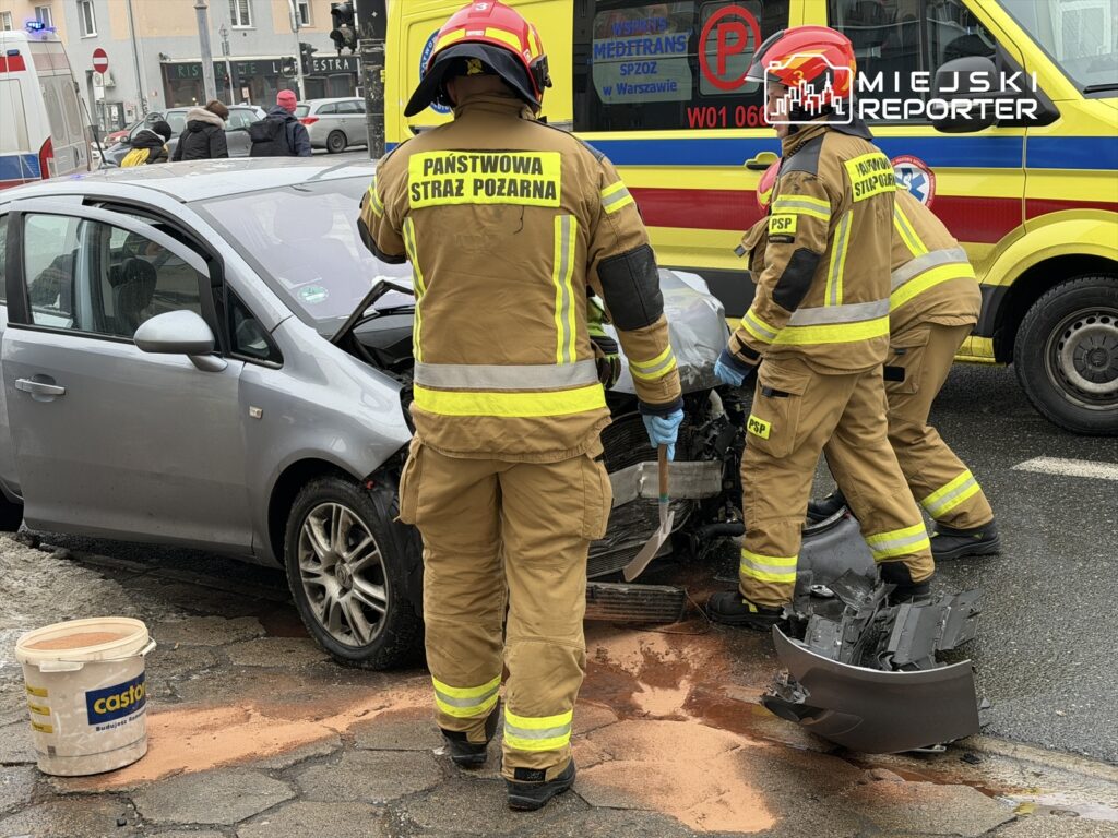 Strażacy w mundurach pracują nad usunięciem wraku samochodu po wypadku drogowym, obok leży plama oleju.