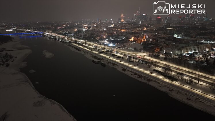 Widok na oświetloną rzekę z nabrzeżem, w tle nowoczesne budynki i świątynie, nocą w zimowej scenerii.