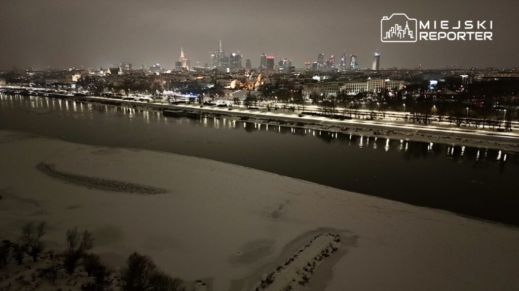 Widok na oświetlone miasto nocą z rzeką pokrytą śniegiem w pierwszym planie, w tle wieżowce i zabytkowe budynki.