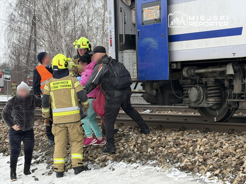 Ratownicy w kaskach pomagają dzieciom wsiąść do pociągu na stacji kolejowej w zimowej scenerii.
