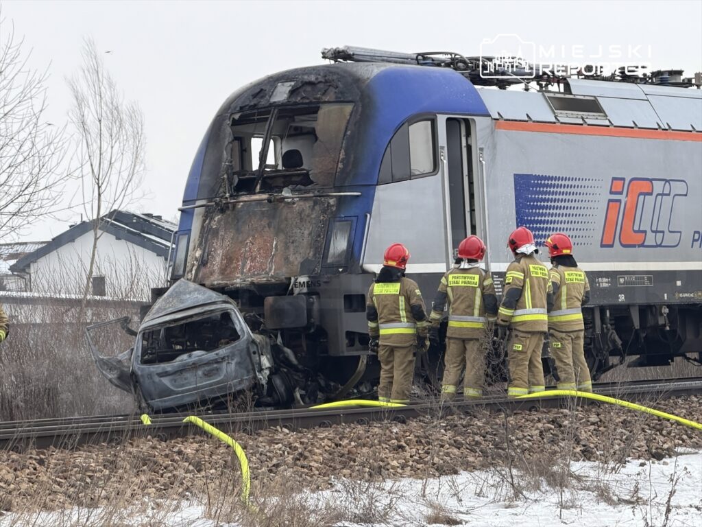 Pojazd kolejowy z uszkodzoną kabiną stoi obok rozbitego samochodu, a strażacy w hełmach badają sytuację.