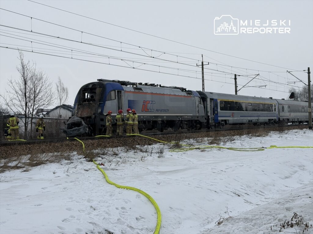 Grupa strażaków w hełmach pracuje przy uszkodzonej lokomotywie na torach w zimowym otoczeniu.
