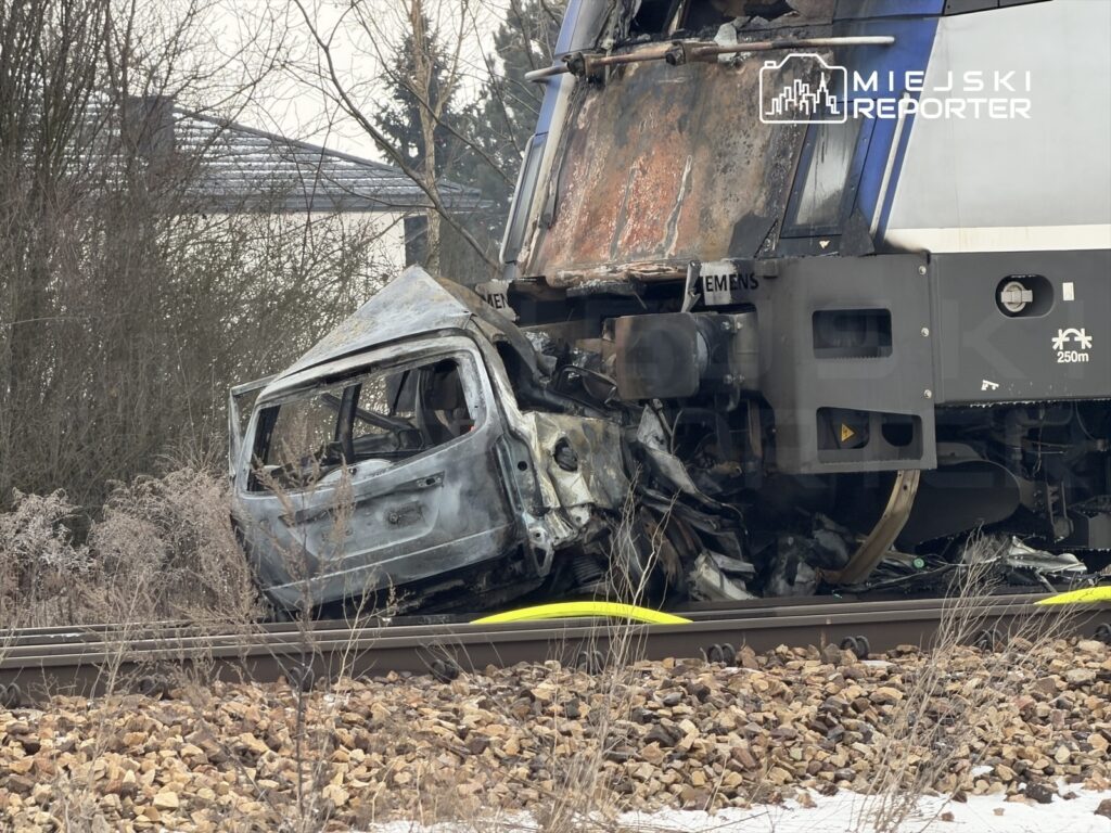 Zderzenie pociągu z samochodem osobowym na torach, z uszkodzoną kabiną pojazdu i pociągiem w tle.