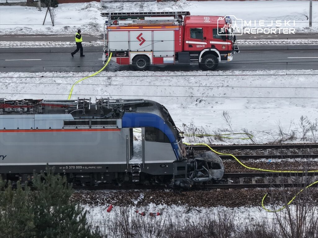 Pociąg towarowy z uszkodzoną lokomotywą stoi na torach, obok niego strażacy rozkładają węże gaśnicze na zaśnieżonym terenie.