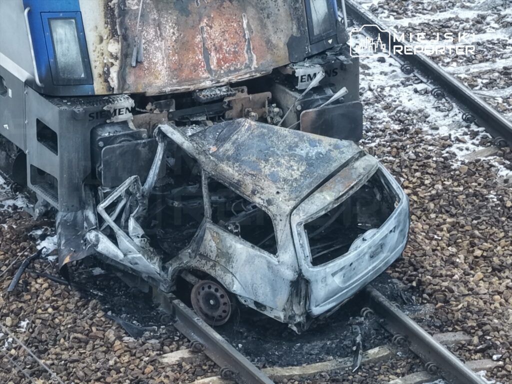 Spalony samochód osobowy leży na torach kolejowych obok uszkodzonego lokomotywy. W tle widoczne kamienie torowiska.